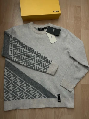 Grå stickad Fendi tröja  - Säljer en snygg ljusgrå stickad tröja från Fendi med ikoniskt grått logomönster på ena ärmen och nedre delen av tröjan. Rund halsringning, ribbade muddar och diskret Fendi-tag med knappdetalj. Perfekt för dig som gillar exklusiv streetstyle.