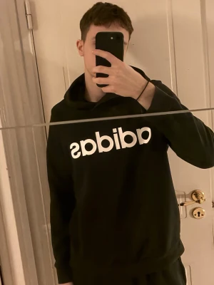 Svart hoodie från Adidas med logga - Snygg svart hoodie från Adidas med stor vit logga framtill. Klassisk modell med huva och ribbade muddar vid ärmslut och nederkant. Perfekt för en avslappnad och sportig stil.