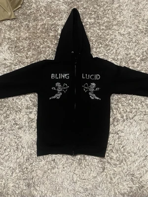 Svart Bling Lucid hoodie med strass och BLING LUCID - Svart hoodie med huva och dragkedja framtill. På bröstet står det 'BLING LUCID' i strass och under texten finns två änglar i strass. Hoodien har fickor framtill och en clean, oversized vibe.