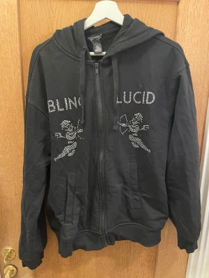 Svart hoodie BLING LUCID med strass - Svart zip-hoodie från BLING LUCID med huva och dragsko. Framsidan har strasstenar som bildar texten 'BLING LUCID' och två änglar. Mjukt material och två fickor framtill. Perfekt för en cool och avslappnad stil.