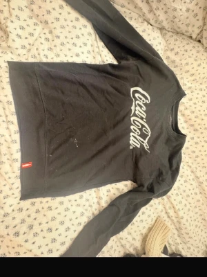 Svart Coca-Cola sweatshirt - Säljer en svart sweatshirt från Coca-Cola med den klassiska loggan tryckt i vitt på bröstet. Tröjan har rund halsringning och långa ärmar. Tillverkad i mjukt bomullsmaterial för en skön och avslappnad look.