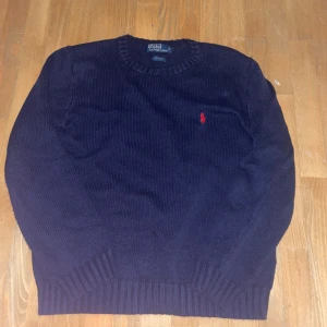 Mörkblå stickad tröja från Polo Ralph Lauren - Mörkblå stickad tröja från Polo Ralph Lauren med rund halsringning och röd broderad logga på bröstet. Tröjan har ribbstickade muddar vid ärmslut och nederkant. Perfekt för en stilren och klassisk look.