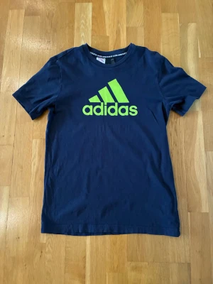 Mörkblå Adidas t-shirt med logga - Snygg mörkblå t-shirt från Adidas med den klassiska loggan i neon-grönt framtill. Tillverkad i mjuk bomull och har en normal passform med korta ärmar. Perfekt för dig som gillar sportig stil och vill sticka ut lite extra.