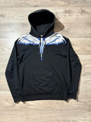Marcelo Burlon - Snygg svart hoodie från Marcelo Burlon med ikoniskt tryck av blå och vita vingar över axlar och bröst. Hoodien har huva med dragsko och en stor magficka framtill. Perfekt för dig som vill sticka ut med en unik och trendig stil.