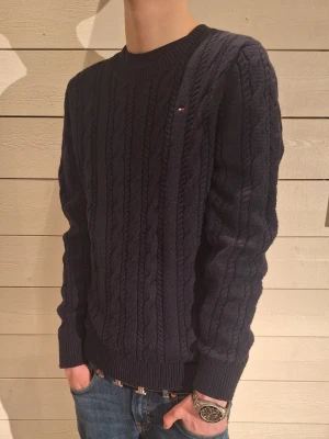 Mörkblå kabelstickad tröja Tommy Hilfiger - Mörkblå kabelstickad tröja från Tommy Hilfiger med rund halsringning och diskret logga på bröstet. Tröjan har ribbade muddar vid ärmslut och nederkant, och är gjord i ett mjukt material som passar perfekt till jeans. Modelen är 172!