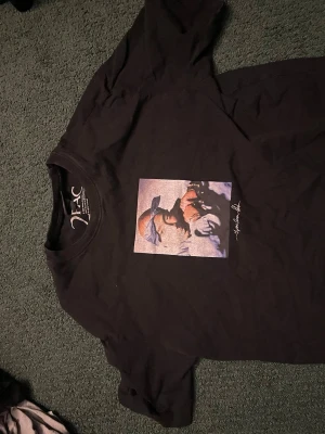 Svart t-shirt med Tupac-tryck - Svart t-shirt från XLC med ett stort färgglatt tryck av Tupac på bröstet. Klassisk rund hals och korta ärmar. Perfekt för dig som gillar streetwear och vill ha en unik look.