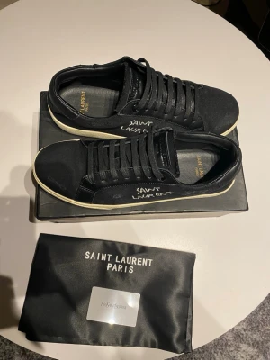 Svarta Saint Laurent sneakers storlek 44,5 - Svarta sneakers från Saint Laurent med vit sula och handskriven logga på sidan. Skorna har snörning och är tillverkade i canvas och skinn. Klassisk låg modell med rund tå och diskreta detaljer. Perfekta för dig som gillar stilrena och exklusiva sneakers.
