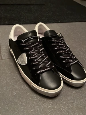 Svarta Philippe Model sneakers - Stilrena svarta sneakers från Philippe Model med vita detaljer och logga på sidan. Skorna har svarta snören med vit text och en vit sula. Tillverkade i skinn för en lyxig känsla och passar perfekt till streetwear eller casual outfits. (Rep)