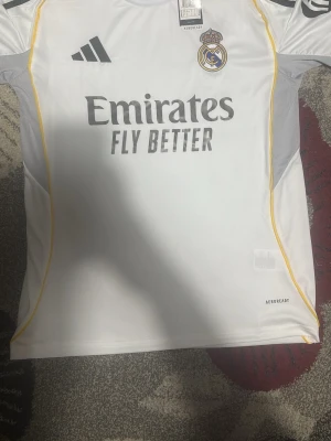 Real Madrid matchtröja Adidas vit - Snygg vit Real Madrid matchtröja från Adidas med klubbmärke och sponsortryck på bröstet. Tröjan har svarta detaljer, gula linjer längs sidorna och är tillverkad i lätt och ventilerande AEROREADY-material. Perfekt för fotbollsträning eller att bära på läktaren.