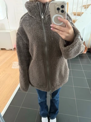 Brun teddyjacka  - Mysig brun teddyjacka med hög krage och hel dragkedja framtill. Jackan har en loose passform och är tillverkad i fluffigt teddy-material som håller dig varm. Perfekt för kyliga dagar och enkel att styla till jeans eller leggings.