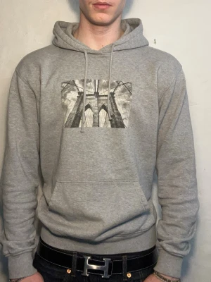 Grå hoodie med brotryck - Snygg grå hoodie med ett svartvitt tryck av en bro på bröstet. Klassisk känguruficka framtill och justerbar huva med dragsko. Tillverkad i mjukt sweatshirtmaterial som är skönt mot huden. Perfekt för dig som gillar streetwear och enkel stil.