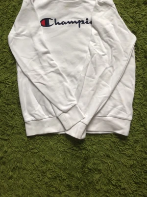 Vit sweatshirt från Champion - Säljer en vit sweatshirt från Champion med det klassiska broderade logotypen i blått, rött och vitt på bröstet. Tröjan har rund hals, långa ärmar och ribbade muddar vid ärmslut och nederkant. Perfekt för en avslappnad och sportig stil.