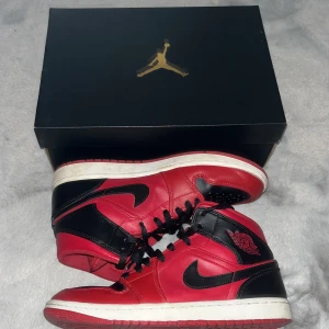 Nike Air Jordan 1 Mid röd och svart - Säljer ett par Nike Air Jordan 1 Mid i klassisk röd och svart färg med röda sulor. Skorna har svart snörning, rund tå och är tillverkade i skinn. Perfekta för dig som gillar streetwear och vill sticka ut med en ikonisk sneaker.