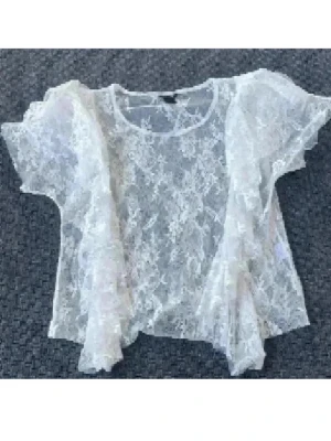 Vit transparent spetsblus med volanger - Säljer en superfin vit transparent topp i spets med korta, vida ärmar och volangdetaljer framtill. Blusen har ett blommigt spetsmönster och rund halsringning. Perfekt för dig som gillar romantisk och trendig stil.