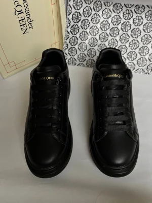 Svarta sneakers från Alexander McQueen - Stilrena helsvarta sneakers från Alexander McQueen med rund tå, platt sula och snörning. Skorna är tillverkade i slätt skinn och har diskret logga i guld på plösen. Perfekta för dig som gillar en clean och modern look.