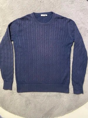 Mörkblå kabelstickad tröja från J.Lindeberg - Snygg mörkblå kabelstickad tröja från J.Lindeberg Tröjan har rund halsringning, långa ärmar och ribbade muddar vid ärmslut och nederkant. Perfekt för dig som gillar klassisk stil med en modern twist.