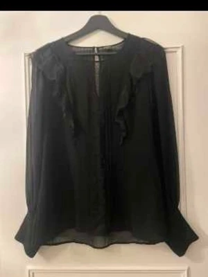 Svart blus med volangdetaljer - Snygg svart blus i transparent material med volangdetaljer vid axlarna och dekorativa knappar vid ärmsluten. Blusen har rund halsringning och långa, puffiga ärmar. Perfekt att styla med jeans eller kjol för en trendig look.