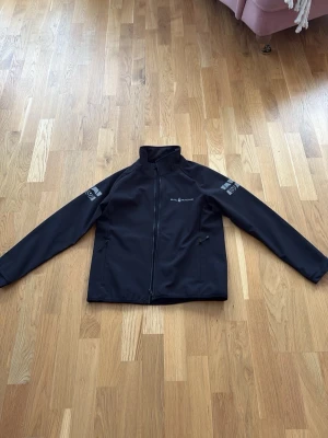 Svart vindjacka från Sail racing - Svart vindjacka från sail racing i storlek 150 med hög krage och dragkedja framtill. Jackan har två sidofickor med dragkedja och tryckta loggor på ärmar och rygg. perfekt för blåsiga dagar. Jag är 167 och jackan är i storlek 150