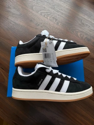 Svarta Adidas Campus sneakers - Klassiska Adidas Campus sneakers i svart mocka med vita detaljer och de ikoniska tre ränderna på sidan. Skorna har vit sula med gummiyttersula och snörning framtill. Perfekta för en clean och tidlös streetstil.