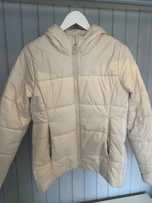 Beige pufferjacka från Adidas -  Beige pufferjacka från Adidas med huva och dragkedja framtill. Jackan har diskret Adidas-logga på bröstet och klassiska ränder längs sidorna. Materialet är polyester. Inte använd mycket alls men lite smink som fasnat som man kan se på bilden, kanske går att få bort men inget jag kan garanter. Sitter bra på mig som har storlek S eller 36-38. 