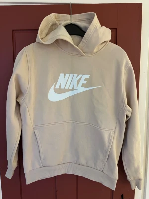 Beige Nike hoodie med stor logga - Snygg beige hoodie från Nike med stor vit logga på bröstet. Tröjan har huva, magficka och ribbade muddar vid ärmslut och nederkant. Insidan är mjuk och fleecefodrad för extra komfort. Perfekt för chill eller streetwear.