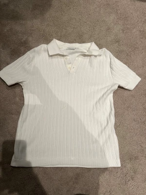 Vit ribbad pikétröja från Kappahl - Vit pikétröja från Kappahl i storlek S. Tröjan har korta ärmar, klassisk krage och knappar framtill. Materialet är ribbad och känns mjukt mot huden, perfekt för vår och sommar. Enkel och stilren design som funkar till många olika looks.