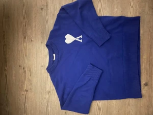 Blå stickad tröja från Ami Paris - Snygg blå stickad tröja från Ami Paris med ett vitt hjärta och bokstaven A på bröstet. Tröjan har ribbade muddar vid ärmslut och nederkant samt rund hals. Perfekt för dig som gillar stilrena detaljer och vill ha något som sticker ut.