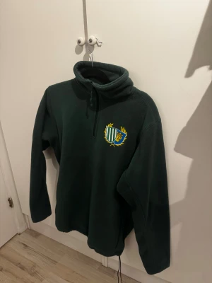 Hammarby fleece tröja - Mörkgrön fleece-tröja med halv dragkedja och hög krage. På bröstet finns ett broderat emblem i grönt, vitt, blått och gult med en krona och sköldar. Tröjan har lång ärm och är perfekt för kyliga dagar. Skön och mjuk känsla tack vare fleecematerialet.