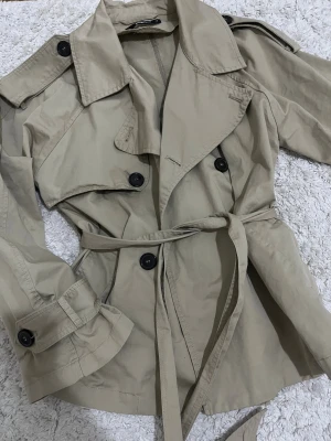 Beige trenchcoat från Gina Tricot XS - Stilren beige trenchcoat från Gina Tricot i storlek XS. Jackan har klassisk krage, knappar, axelklaffar och bälte i midjan. Tillverkad i bomullsmaterial och perfekt för vår och höst. Snygg och tidlös modell med långa ärmar och fina detaljer. Helt slutsåld! 