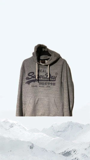 Grå hoodie från Superdry med tryck - Snygg grå hoodie från Superdry med stort mörkblått logotryck på bröstet och japanska tecken. Tröjan har huva med vita snören och en klassisk känguruficka framtill. Perfekt för chill dagar eller när du vill ha en sportig look.