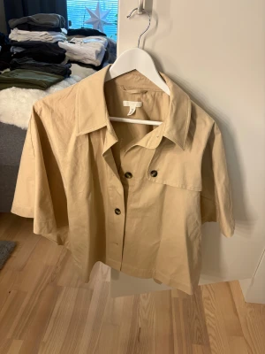 Beige trenchcape från H&M - Snygg beige trenchcape från H&M med korta ärmar och oversized passform. Skjortan har klassisk krage, stora knappar framtill och en cool detalj med extra lager på ryggen. Perfekt att slänga över en hoodie för en chill look.