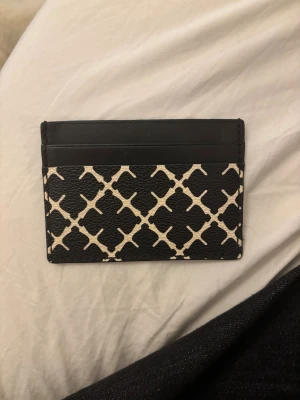 Marlene Birger plånbok - Snygg och stilren kortplånbok i svart och beige med grafiskt mönster. Plånboken är tillverkad i präglat skinn och har flera fack för kort på ena sidan. Perfekt för dig som vill ha en smidig och trendig accessoar i fickan.