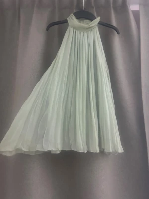 Ljusgrön blus i chiffong - Superfin ljusgrön blus i luftig chiffong med plisserad design och hög hals. Blusen är ärmlös och har en flowy, A-linjeformad siluett som ger en riktigt drömmig känsla. Perfekt för dig som vill sticka ut med en elegant och fräsch look.