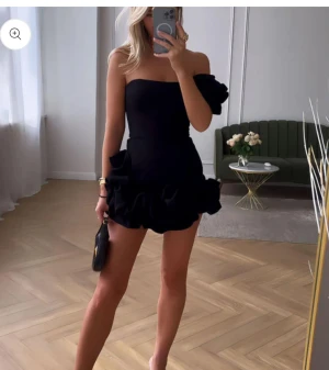 Klänning - Supersnygg svart kort klänning med volangdetaljer och axelbandslös design. En axel har en extra volang för en cool asymmetrisk look. Perfekt för dig som vill sticka ut med en trendig och elegant stil. Storlek M har ej använt. Passar inte mig trodde den va liten i storlek.