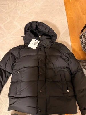 Svart Moncler dunjacka med huva helt ny - Svart dunjacka från Moncler med klassisk puffer-design, huva och stora fickor framtill. Jackan har Moncler-logga på ärmen och stängs med både knappar och dragkedja. Perfekt för kalla dagar och riktigt snygg streetstil.