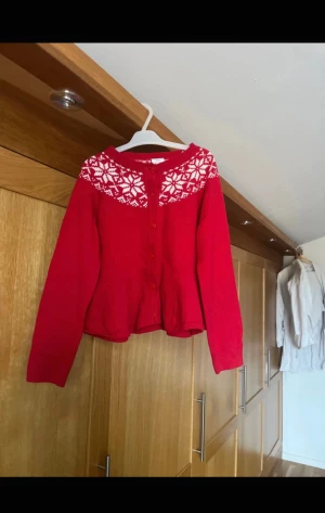 Röd kofta från Polarn O. Pyret - Superfin röd kofta från Polarn O. Pyret med vita snöflingemönster runt halsen. Koftan har rund hals, långa ärmar och knappar framtill. Perfekt för vintern och julen, stickad i mjukt material som håller dig varm och snygg.alldrig anndvänd