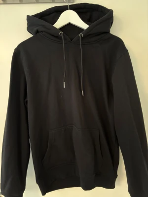 Svart hoodie från Urban Classics M - Svart hoodie från Urban Classics i storlek M. Klassisk modell med huva och snörning, stor magficka framtill och mjukt material i bomull och polyester. Perfekt för chill dagar och enkel att matcha med jeans eller joggers.