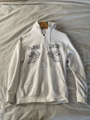 Vit hoodie Bling Lucid med änglar - Vit hoodie från Bling Lucid med dragkedja och huva. Framsidan har två svarta änglamotiv och texten 'BLING LUCID' i glittriga stenar. Utrustad med kängurufickor och snörning vid huvan. Perfekt för dig som gillar streetwear med unik design.