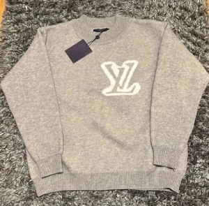 Grå stickad Louis Vuitton tröja - Snygg grå stickad tröja från Louis Vuitton med stort vitt LV-logo framtill. Tröjan har rund hals, ribbade muddar och är långärmad. Perfekt för dig som gillar exklusiva märken och vill ha en stilren look.