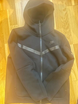 Svart Nike Tech Fleece jacka - Nike Tech Fleece jacka i svart med hel dragkedja och huva. Jackan har en diskret logga på bröstet, svarta detaljer och en diagonal rand över bröstet. Ärmarna är långa och det finns en ficka med dragkedja på ena ärmen. Perfekt för en sportig och clean look.