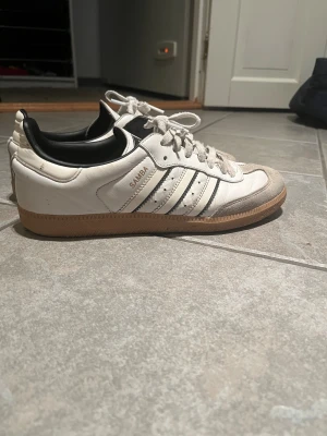 Adidas Samba vita sneakers - Nypris 1299kr, köpa i våras. Klassiska Adidas Samba sneakers i vitt skinn med svarta detaljer och gummisula. Skorna har de ikoniska tre ränderna på sidan, svart hälkappa och grå mockadetaljer vid tån. Perfekta för dig som gillar retrostil och streetwear.