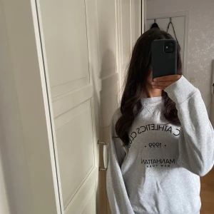 Grå NYC Athletics Club sweatshirt - Snygg grå sweatshirt från NYC Athletics Club med svart tryck på framsidan: 'NYC ATHLETICS CLUB 1999 MANHATTAN NEW YORK'. Tröjan har rund hals, ribbade muddar och är långärmad. Perfekt för chill dagar eller när du vill ha en sportig vibe.