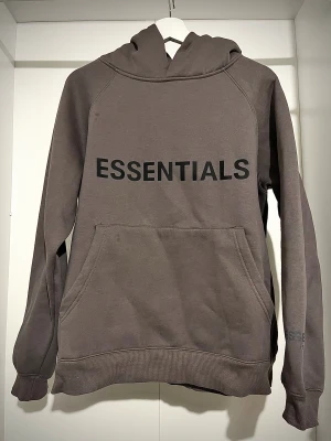  Essentials hoodie  - Säljer en mörkgrå hoodie från Fear of God Essentials med stor svart logga på bröstet och mindre tryck på ärmen. Tröjan har huva, känguruficka och är långärmad. Perfekt för dig som gillar streetwear och vill ha en clean look.