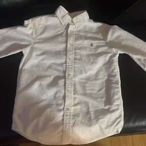 Vit skjorta från Ralph Lauren - Vit klassisk Oxford skjorta från Ralph Lauren med broderad logga på bröstet. Skjortan har lång ärm, knappar hela vägen och en snygg krage. Tillverkad i mjuk bomull och har en clean, stilren look som funkar till många olika outfits.