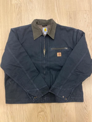 Carhartt workwear jacka M - Mörkblå (heavy water wash) workwear jacka från Carhartt med kontrasterande brun krage och rutigt innerfoder. Jackan har dragkedja framtill, bröstficka med dragkedja och klassisk Carhartt-logga. Tillverkad i slitstark bomull, perfekt för en streetwear-look.