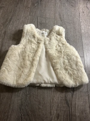 Krämvit pälsväst från H&M barn - Supermjuk och fluffig krämvit pälsväst från H&M för barn. Västen har ett lent foder och stängs med en diskret knapp framtill. Perfekt för att ge outfiten en mysig och trendig touch under kyligare dagar.