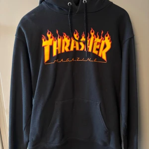 Svart Thrasher hoodie med flames - Svart hoodie från Thrasher med ikoniskt flames-tryck i orange och gult över bröstet. Tröjan har huva med snörning, stor magficka och är tillverkad i mjuk bomull och polyester. Perfekt för dig som gillar streetwear och skatekultur. Anvönd några gånger men fortfarande i mycket bra skick
