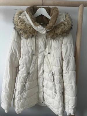 Vit pufferjacka med fuskpäls från Zara - Snygg vit pufferjacka från Zara i storlek XL. Jackan har en mysig huva med beige fuskpäls och dragkedjor på fickorna. Perfekt för kalla dagar med sin quiltade design och fluffiga känsla. Stängs med dragkedja framtill och har långa ärmar.