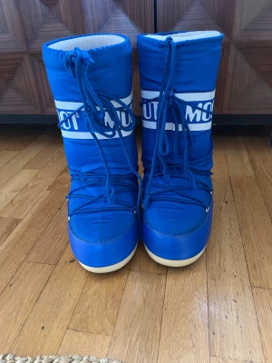 Blå Moon Boots med snörning - Säljer ett par ikoniska blå Moon Boots med vit logga och snörning framtill. Skorna har en rund tå, platt sula och är fodrade med mjukt, ljust material på insidan. Perfekta för vinter och snö, med slitstark syntet på utsidan och greppvänlig gummisula. Knappast använda, perfekt skick!!