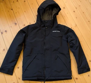 Svart vinterjacka Burton Dugout M barn 140/146 - aldrig använd! - Helt ny, aldrig använd, svart vinterjacka från Burton med huva och dragkedja. Modell: Dugout, stl 140/146. Jackan har flera fickor med dragkedja och en quiltad insida i grått. Köpt för 1599 kr för två år sedan - aldrig använd!
